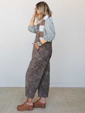A relaxed side-profile shot of a model in acid-washed brown floral-print overalls layered over a striped shirt, styled with wood-sole clogs and a stack of turquoise and pearl bracelets — the kind of effortless outfit that looks like it came together