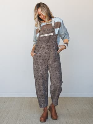 A relaxed full-body shot of floral-printed brown overalls layered over a color-block long-sleeve top, styled with clogs and stacked bracelets — the kind of outfit that feels effortless but totally thought-out.