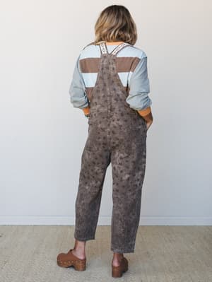 A back-facing shot of a relaxed floral-print overall styled over a striped colorblock long-sleeve top, finished with brown studded clogs — the kind of layered, lived-in look that feels intentional without trying too hard.