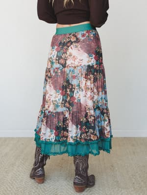 A back-view shot of a tiered patchwork floral maxi skirt styled with a dark brown cropped sweater and brown cowboy boots — the kind of outfit that looks like it was pulled together effortlessly but definitely wasn't.