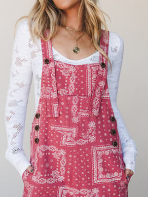 A bandana-print overall dress in a warm raspberry pink layered over a white lace long-sleeve top, styled with gold layered necklaces — the kind of outfit that feels effortless but totally intentional.