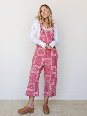 A bandana-print cropped overall in a warm rosy pink, layered over a white eyelet long-sleeve and finished with tan clogs — the kind of outfit that looks like it happened naturally but absolutely did not.