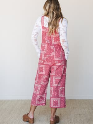 A model shot from behind showing wide-leg cropped overalls in a warm rose-pink bandana patchwork print, layered over a white lace long-sleeve top and styled with brown wooden clogs — casual, a little nostalgic, and very easy to live in.