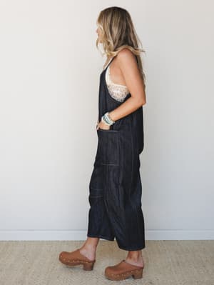 A relaxed dark denim jumpsuit styled with a cream lace bralette and brown clogs — the kind of effortless outfit that looks like you tried just enough.