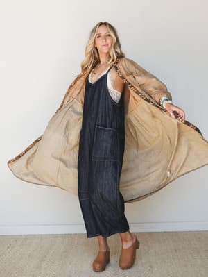 A relaxed, layered look — dark denim midi jumpsuit worn over a white lace bralette, paired with a tan quilted duster coat with leopard trim and brown clogs, styled against a clean white wall with an easy, confident stance.