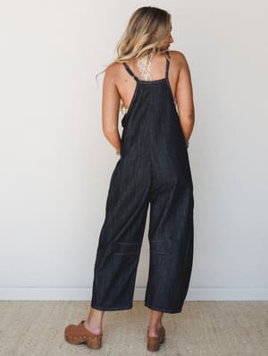 A relaxed dark denim overall jumpsuit shot from the back, styled with a lace bralette and wood-heeled clogs — the kind of effortless outfit that looks like you tried just enough.