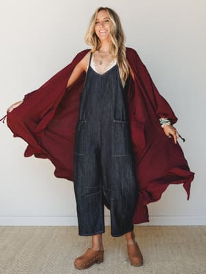 A smiling model opens a flowing burgundy duster wide to reveal a dark denim wide-leg jumpsuit layered over a lace cami, styled with tan clogs and stacked bracelets — the kind of outfit that looks effortless but clearly wasn't an accident.