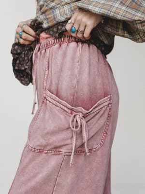 A close-up detail shot of acid-washed dusty rose wide-leg pants with a tied bow pocket, layered under a plaid flannel and a dark floral top, styled with stacked silver and turquoise rings for a collected boho feel.