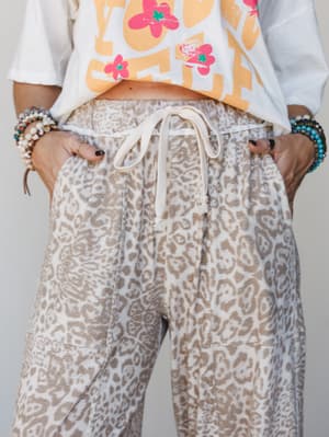 A close-up torso shot of leopard-print joggers in a soft mocha and cream colorway, styled with a cropped graphic tee and stacked bead bracelets — the kind of outfit that feels effortless without trying.