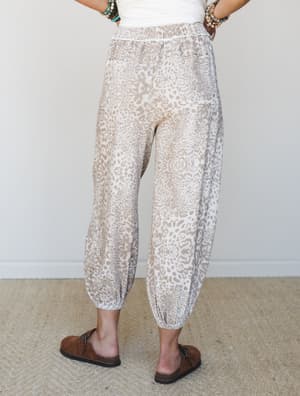 A back-view shot of relaxed leopard-print joggers in a soft mocha and cream colorway, styled with a white tank and Birkenstock-style sandals — the kind of pants that make a coffee run feel like a personality.