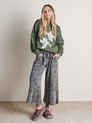 Woman wearing a green sweater with a floral design and gray floral pants, standing against a plain background.