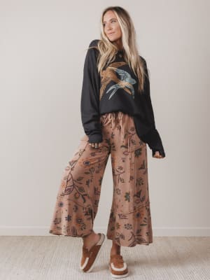 Woman wearing a black hoodie with floral design and brown floral pants against a plain background