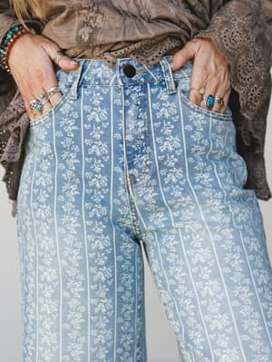A close-up detail shot of wide-leg denim jeans in a soft washed blue, printed with an all-over white floral and vertical stripe pattern — hands tucked into front pockets, layered with silver and turquoise rings and a colorful beaded bracelet, styled