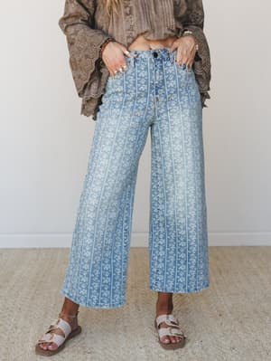 Wide-leg floral print jeans in light washed denim styled with a mocha eyelet blouse and buckle sandals — the kind of outfit that makes a Tuesday feel like something worth dressing for.
