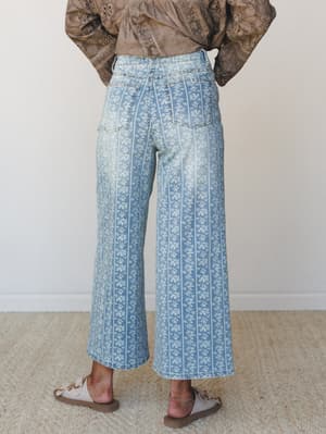 Shot from behind, this outfit pairs wide-leg floral-stripe denim jeans with a mocha eyelet blouse and neutral sandals — a lived-in boho combo that feels like a Sunday well spent.