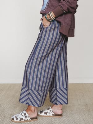 A side-profile shot of wide-leg striped pants in navy, grey, and rose paired with a washed burgundy graphic sweatshirt, stacked bead bracelets, and cow-print slide sandals — the kind of effortlessly collected outfit that looks thrown together in the