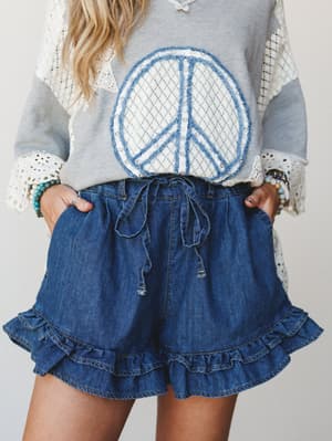 A close-up torso shot of ruffle-hem denim shorts paired with a grey graphic sweatshirt featuring a peace sign appliqué and lace bell sleeves — the kind of outfit that feels effortlessly nostalgic and a little bit fun.