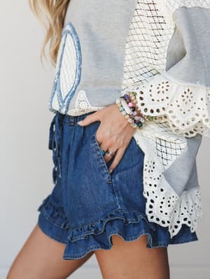 A close-up detail shot of ruffle-hem denim shorts paired with a mixed-media lace and eyelet top, styled with stacked beaded bracelets and rings — the kind of outfit that feels put-together without trying too hard.