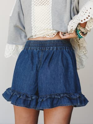 A close-up mid-shot of ruffle-hem denim shorts styled with a gray top layered over a cream lace piece, accessorized with a stack of beaded bracelets — easy, boho, and just the right amount of playful.