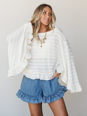 A cream open-knit sweater with dramatic ruffle-trimmed bell sleeves is paired with washed denim ruffle-hem shorts — the kind of outfit that looks effortless but is clearly doing a lot of good work.
