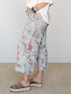 A side-profile shot of wide-leg cropped pants in a soft grey waffle knit with an all-over vintage floral print, styled with a white graphic tee, stacked beaded bracelets, and leopard print platform sandals — relaxed and a little lived-in in the best