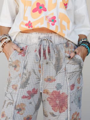 A close-up of floral waffle-knit drawstring pants in faded grey paired with a graphic crop tee and stacked beaded bracelets — the kind of outfit that looks effortless but is absolutely a whole vibe.