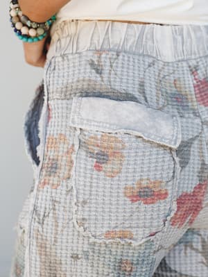 A close-up detail shot of soft grey waffle-knit pants with a faded floral print and raw-edge patchwork pocket, styled with a white top and a stack of beaded boho bracelets.