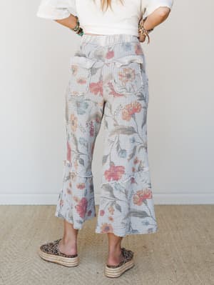 A back-view shot of cropped flare pants in a soft grey waffle knit with a watercolor floral print, styled with a white crop tee, stacked bracelets, and leopard espadrille slides — the kind of outfit that looks effortless but was absolutely thought ab