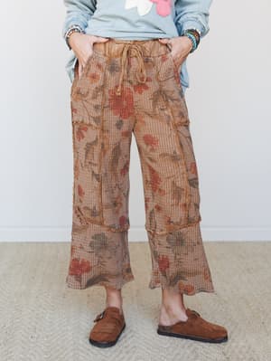 A cropped wide-leg pant in a warm mocha waffle-knit with an earthy floral print, styled casually with a sage appliqué sweatshirt, beaded bracelets, and suede clogs — the kind of outfit that looks effortless but was definitely thought about.