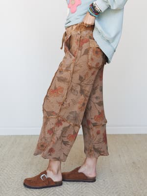 A side-profile shot of wide-leg cropped pants in a warm mocha waffle-knit fabric with a faded floral print, styled with a light blue graphic sweatshirt, stacked beaded bracelets, and brown suede clogs — earthy, lived-in, and effortlessly personal.
