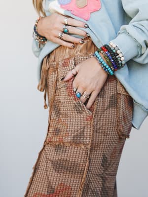 A close-up detail shot focused on stacked beaded bracelets and layered silver rings against a textured waffle-knit pant with frayed edges, paired with a light blue floral appliqué sweatshirt — the kind of jewelry stack that takes years to collect and