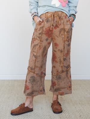 Cropped wide-leg pants in a warm mocha waffle knit with an earthy floral print, styled with a dusty blue floral appliqué sweatshirt, stacked beaded bracelets, and brown suede clogs — the kind of outfit that looks like you tried just enough.