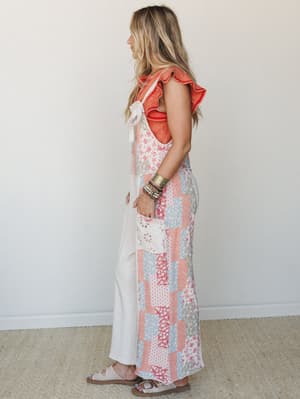 A side-profile shot of a patchwork maxi jumpsuit in soft coral, dusty blue, and cream florals, layered over a ruffle-sleeve coral knit top and finished with stacked bracelets and slide sandals — the kind of outfit that looks like it came together eff