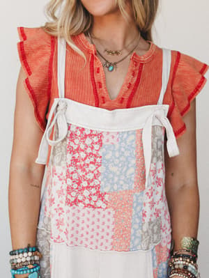 A layered boho look featuring a coral flutter-sleeve top peeking out beneath a patchwork floral bib-style jumpsuit in cream — stacked with turquoise bracelets and layered necklaces for that collected, effortlessly personal feel.