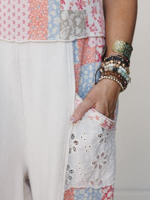 A close-up detail shot of an ecru jumpsuit with an eyelet lace pocket and patchwork floral top panel, styled with a layered stack of mixed boho bracelets — the kind of outfit detail that makes someone stop scrolling.