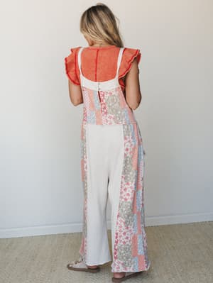 Shot from behind, a model wears a wide-leg patchwork jumpsuit in cream with floral panels in coral, dusty blue, and gray, layered over a coral ruffle-sleeve top — the back button detail and relaxed silhouette giving it a vintage quilt-meets-summer-af