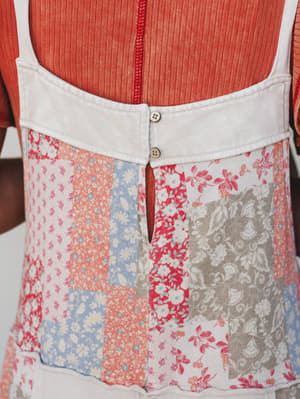 A close-up detail shot of a patchwork floral skirt or jumpsuit layered over a rust ribbed top, showing the back waistband with button closures and a mix of vintage-feel floral prints in coral, dusty blue, red, and taupe.