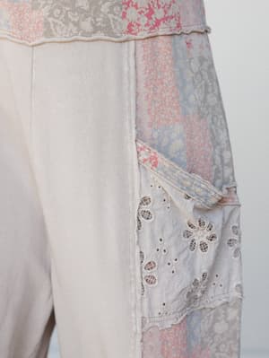 A close-up detail shot of a patchwork jumpsuit panel featuring eyelet lace pocket, mixed floral fabric panels in soft pinks and blues, and raw-edge seam construction on a cream base.