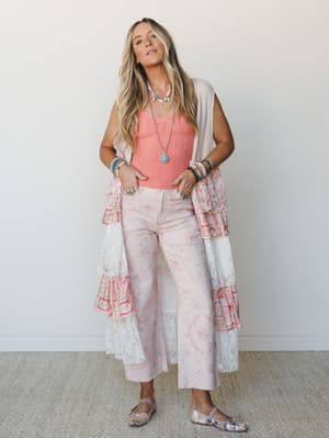 A full-body lifestyle shot of a model layered in soft blush and coral — floral-printed wide-leg jeans, a textured coral tank, and a floor-length lace and mixed-print duster vest, finished with stacked turquoise jewelry and metallic ballet flats for a