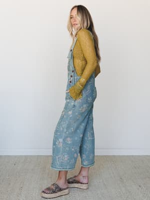 A floral-printed light denim overall styled over a golden olive lace long-sleeve top, shot in profile against a clean white studio wall — the kind of outfit that looks like it was pulled together effortlessly but absolutely was not.