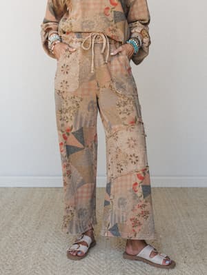 A patchwork-print wide-leg pants in warm latte tones styled with a matching printed top, stacked beaded bracelets, and white buckle sandals — the kind of outfit that looks like it came together effortlessly but definitely did not.