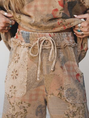 A close-up detail shot of mixed-print drawstring pants in a warm latte wash, featuring an eyelet lace pocket patch and a matching printed top — the kind of lived-in set that looks like it was made from vintage fabric scraps in the best possible way.