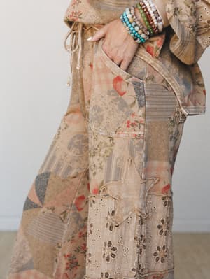 A close-up detail shot of wide-leg patchwork pants in warm latte tones, layered with mismatched florals, stripes, and eyelet panels — worn with a stacked beaded bracelet collection that feels like a weekend flea market find in the best way.