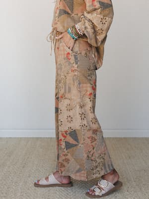A side-profile shot of a patchwork wide-leg pant in warm latte tones, styled with a matching patchwork top, stacked beaded bracelets, and white buckle sandals — the kind of outfit that looks like it has a whole story behind it.