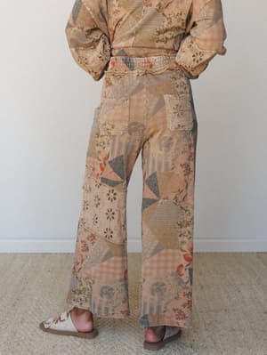 A back-facing model shows off wide-leg pants in a warm latte-toned patchwork print — floral, plaid, and geometric blocks layered together with eyelet embroidery details, styled as a matching set with a coordinating top and worn with casual sandals.