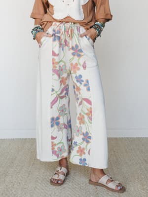 Wide-leg floral pants in cream with a retro-inspired pastel print styled with a caramel linen top, crochet white crop, stacked bracelets, and buckle sandals — the kind of outfit that looks like it was thrown together perfectly.