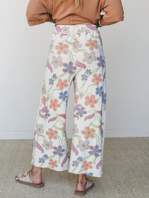 A back-view shot of wide-leg floral pants in a creamy base with a retro-inspired daisy print in peach, periwinkle, soft blue, and pink, styled with a caramel cropped sweatshirt and neutral slide sandals.