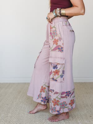 A side-profile shot of wide-leg cargo pants in washed mauve, covered in bold retro floral panels at the hip and hem — paired with a burgundy ribbed tank and stacked boho jewelry for an effortlessly expressive, free-spirited look.