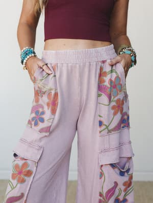 A close-up mid-shot of wide-leg cargo-style pants in washed mauve, covered in bold retro floral print panels, styled with a cropped burgundy tank and stacked beaded bracelets — relaxed, expressive, and full of personality.