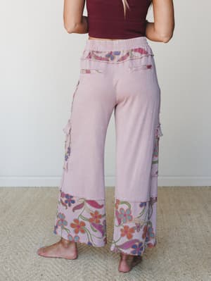 A back-view shot of wide-leg floral pants in washed mauve, styled with a dark burgundy crop top — the mixed-fabric patchwork and bold botanical print at the hem give them that perfectly lived-in, free-spirited feel.
